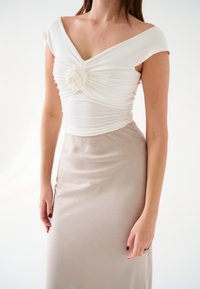 Abito bianco con scollo drappeggiato, caratterizzato da un accento floreale al centro. La gonna è di un beige uniforme con una sottile texture.