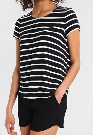 Kobieta ubrana w krótki rękaw w czarno-białe poziome paski i czarne szorty, z jedną ręką w kieszeni, widoczna od szyi do połowy uda.