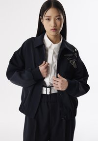 Veste de bombardier marine avec des poignets côtelés, fermeture éclair et accents en métal décoratifs. Portée par-dessus une chemise blanche à boutons et un pantalon noir ceinturé.