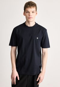 Σκούρο μπλε μπλουζάκι με στρογγυλή λαιμόκοψη, φτιαγμένο από απαλό βαμβάκι, με κοντά μανίκια και μικρό, κυκλικό λογότυπο στο αριστερό στήθος.