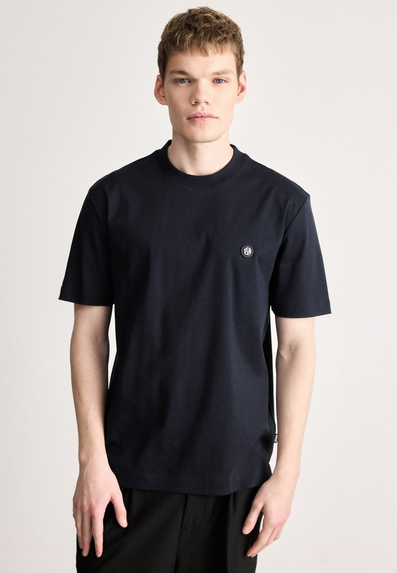 Σκούρο μπλε μπλουζάκι με στρογγυλή λαιμόκοψη, φτιαγμένο από απαλό βαμβάκι, με κοντά μανίκια και μικρό, κυκλικό λογότυπο στο αριστερό στήθος.