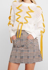 Suéter blanco de punto con bandas texturizadas amarillas, combinado con una falda corta de cuadros marrón y negro que cuenta con bolsillos delanteros.