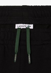 Schwarzer Joggingbund mit einem weißen Lacoste-Label. Grüne Kordeln mit strukturiertem Gewebe, die in metallischen Spitzen enden. Einfaches, funktionales Design.