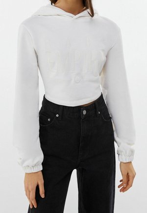 Osoba w białej krótkiej bluzie z bufkami na rękawach i czarnych, wysokich jeansach, stojąca z rękami swobodnie opuszczonymi wzdłuż ciała.