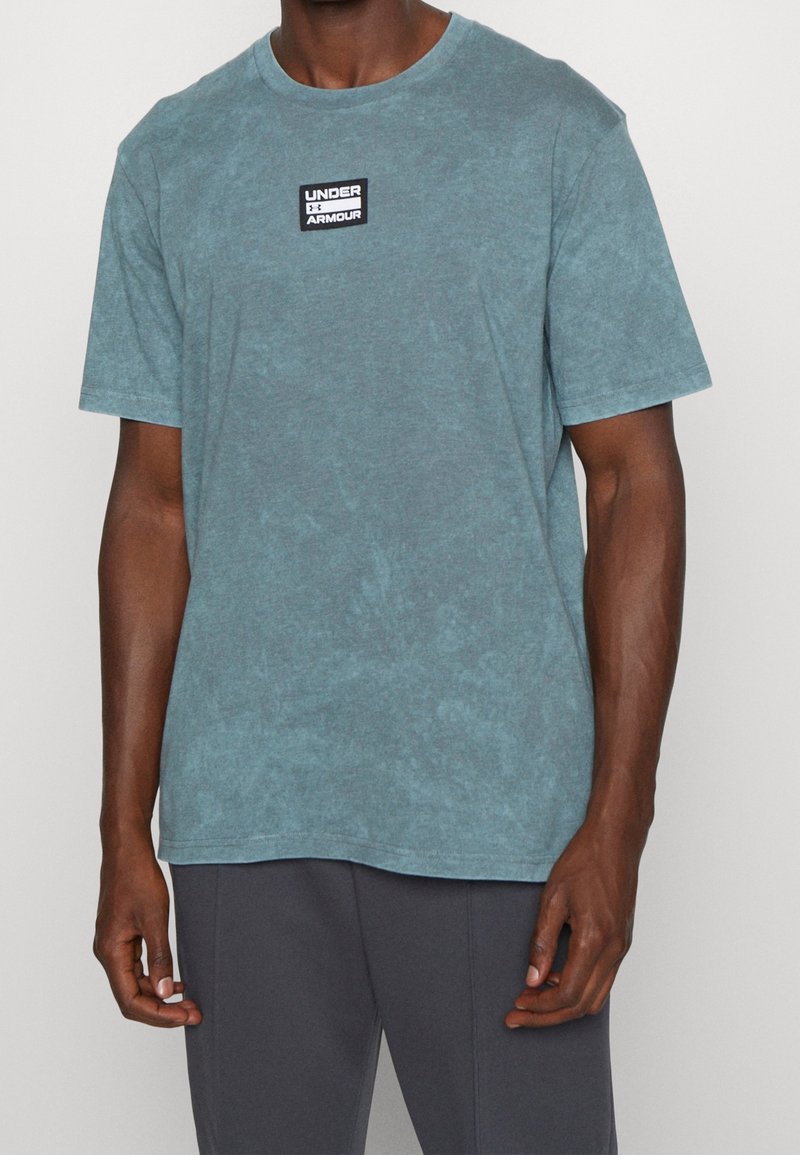 Camiseta de algodón teal con cuello redondo, mangas cortas y una etiqueta negra con "UNDER ARMOUR" en el pecho. Acabado ligeramente texturizado.