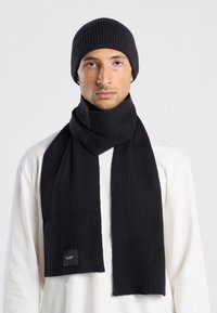 Conjunto de gorro y bufanda de punto acanalado negro, con una textura suave, que presenta una etiqueta cosida en la bufanda; usado sobre una camiseta de manga larga de color claro.