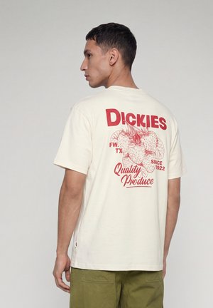 Mann trägt ein cremefarbenes T-Shirt mit roten Erdbeeren und "Dickies Quality Produce FW TX Since 1922" auf dem Rücken, olivgrüne Hose.