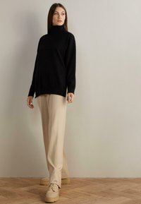 Svart polotröja, löst sittande, långa ärmar, kombinerad med beige vida byxor och ljusbeiga skor med tjocka sulor på träplankgolv.