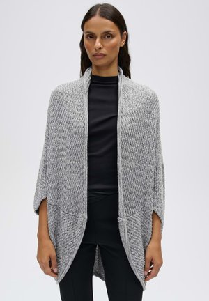 Vrouw draagt een grijze gebreide sjaal over een zwart topje en zwarte broek, staand tegen een effen lichte achtergrond.