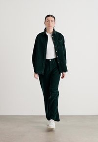Dunkelgrühecordhemd mit Knöpfen, kombiniert mit passenden Cordhosen, getragen über einem weißen, enganliegenden T-Shirt, ergänzt durch weiße Sneakers.