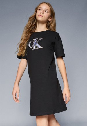 Melnā kokvilnas kleita ar īsām piedurknēm, kurai priekšā ir liels metāliski zils un sudrabs "CK" logo. Vienkārša, brīva piegriezuma.