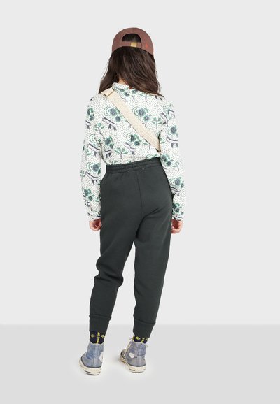 Grønne sweatpants med elastiske manchetter, mønstret langærmet skjorte med blomsterdesign, og beige skulderrem. Brun kasket og blå sko.