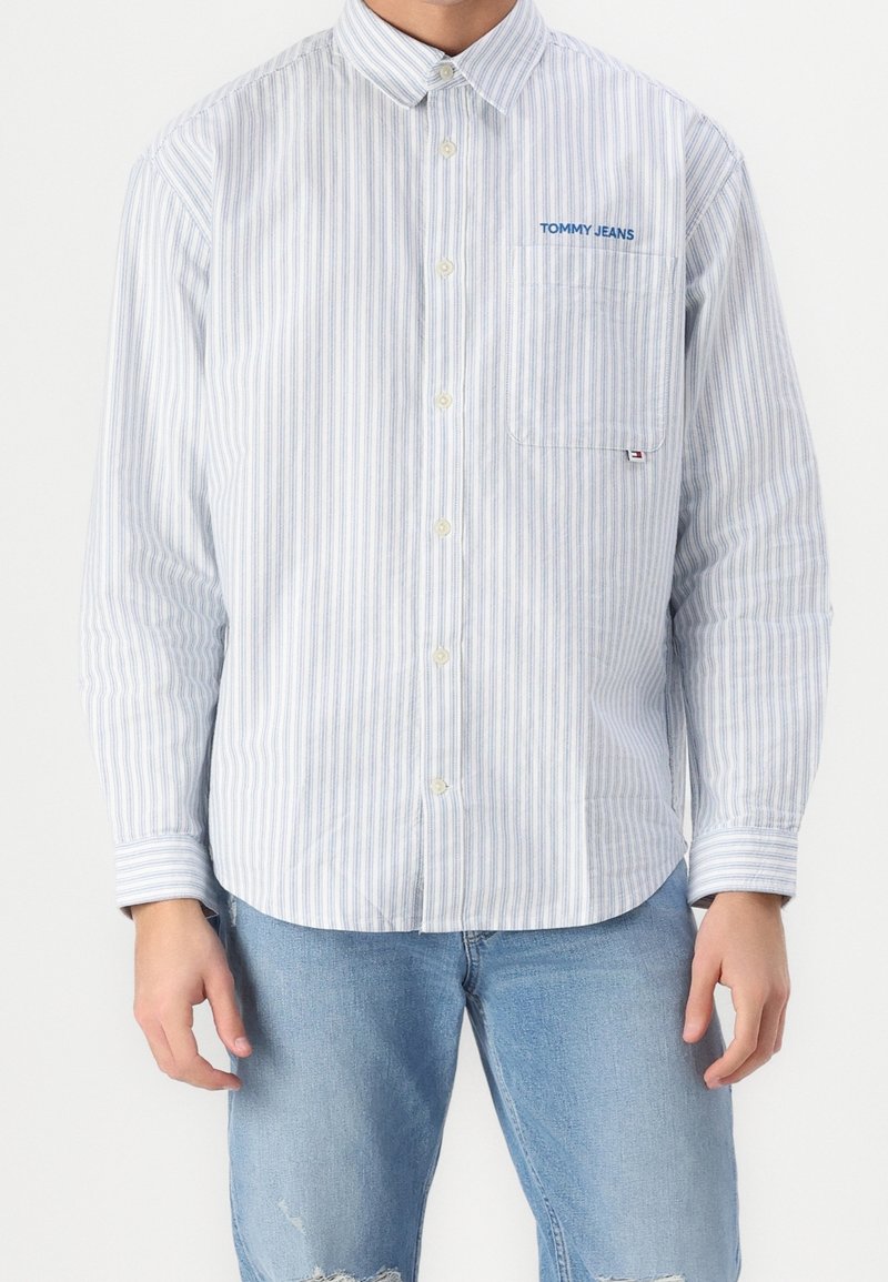 Homme portant une chemise boutonnée à rayures blanches et bleu clair avec le logo "Tommy Jeans" sur la poche, associée à un jean bleu clair déchiré.