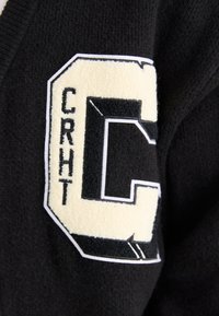 Čierna tkanina s textúrovaným smotanovým filcovým písmom "C" a vyšitými detailmi "CRHT". Dizajn má športový, universitný štýlový estetický vzhľad.