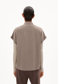 Kortärmad, ljusbrun skjorta med en subtil lyster, med krage och en rundad nederkant, lager på lager över en texturerad, krämfärgad långärmad topp.