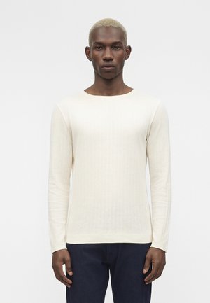 Czarnoskóry mężczyzna z krótkimi, wybielonymi blond włosami, ubrany w dopasowany, kremowy, prążkowany sweter z długim rękawem i ciemne spodnie na jednolitym tle.