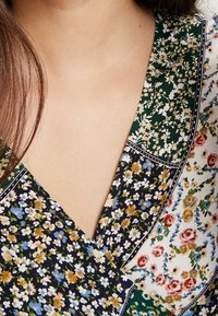 Vestido envolvente floral con un escote en V profundo. Presenta una mezcla de estampados en azul marino, verde y blanco con pequeñas flores y acentos de ribete oscuro.