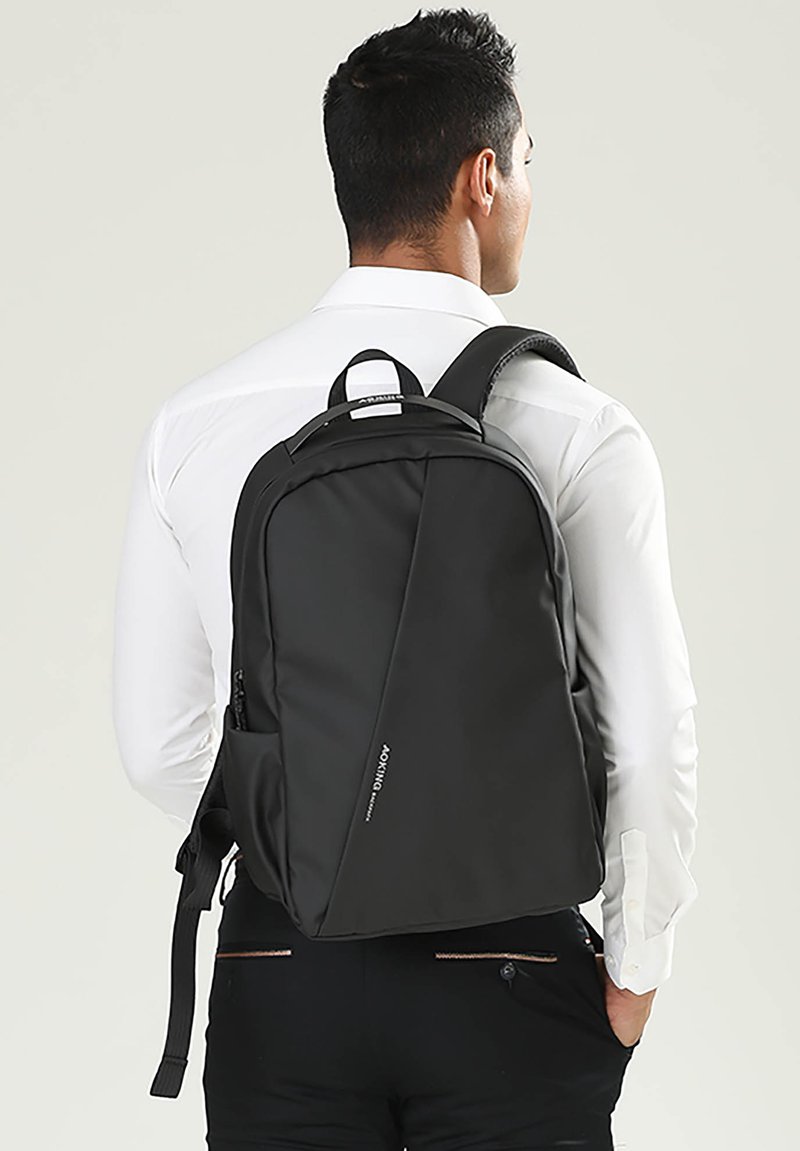 Un sac à dos noir au design élégant, fabriqué en matériau lisse. Il possède des bretelles réglables, une poche latérale et un logo discret à l'avant.