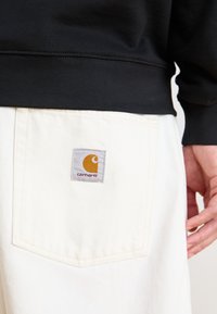 Zwarte sweatshirt rand overlapt met witte denim broek, met een rechthoekig grijs label met oranje logo gestikt op de achterzak.