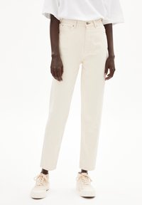 Person med off-white högmidjade raka jeans, vit oversized t-shirt och krämfärgade snörskor står på vit bakgrund.