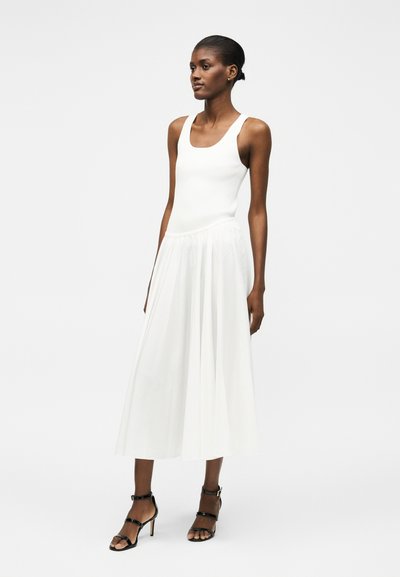 Γυναίκα με λευκό αμάνικο φόρεμα και μαύρα ψηλοτάκουνα σανδάλια στέκεται μπροστά σε απλό φωτεινό φόντο.