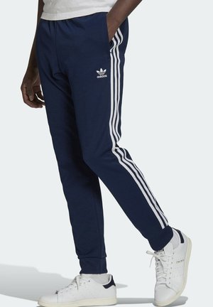 Person trägt marineblaue Adidas-Trainingshose mit weißen drei Streifen und weißen Stan Smith Sneakern, steht mit einer Hand in der Tasche.