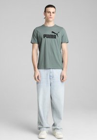 Vihreä puuvillapaita, jossa on musta Puma-logo, yhdistettynä vaaleansinisiin löysiin farkkuihin ja valkoisiin tennareihin. Yksinkertainen, rento asuste.