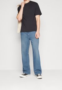 Man med svart t-shirt, ljusblå jeans och svarta låga sneakers, håller en beige jacka över axeln mot vit bakgrund.