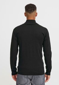 En person med kort, lockigt hår som vänder ryggen mot kameran, iklädd en svart polotröja och grå byxor, mot en helt vit bakgrund.