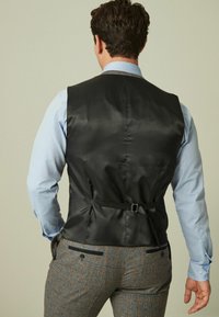 Mann in hellblauem Hemd, schwarzem verstellbarem Rückensack und braunen karrierten Hosen, steht mit den Händen in der Nähe der Taschen und hat seinen Rücken zugewandt.
