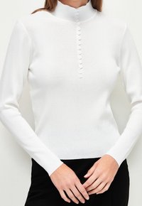 Haut blanc à manches longues avec un col montant, doté d'une patte de boutonnage ornée de petits boutons sur le devant. Texture douce et côtelée avec une coupe ajustée.