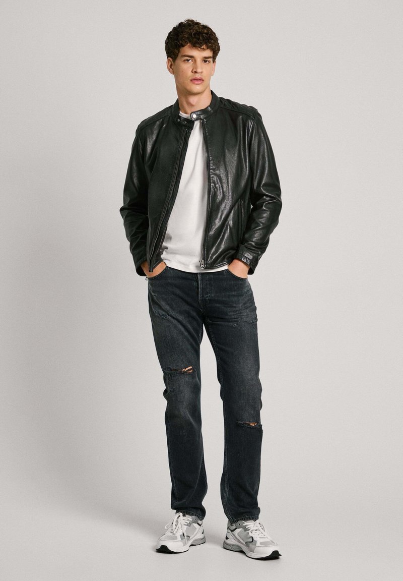 Chaqueta de cuero negra con botón a presión, camiseta blanca, jeans oscuros rasgados y zapatillas gris. El modelo se sostiene con confianza con las manos en los bolsillos.