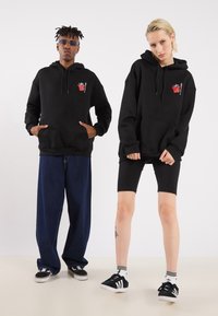 Black hooded sweatshirt with front pocket, worn by two models. One wears loose denim pants, the other black cycling shorts and sneakers.
