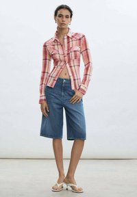 Red and white plaid long-sleeve shirt, cropped style with one chest pocket, paired with blue denim knee-length shorts and white sandals.