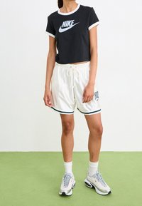 Persona vestida con un crop top negro de Nike, pantalones cortos blancos de Nike con cordón, calcetines blancos y zapatillas blancas de Nike de pie sobre un suelo verde.