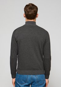 Suéter gris de punto con cuello alto y dobladillo acanalado. Costuras visibles y mangas largas. Combinado con jeans azules, mostrando un estilo casual.