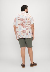 Chemise à manches courtes en tissu léger avec un motif comportant des carrés rouges, oranges et bleus ; associée à un short vert olive et des baskets brunes.