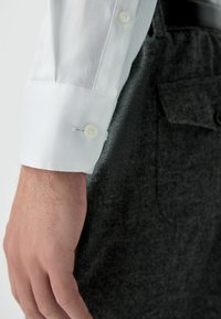 Valkoinen teksturoitu paita hihan päällinen, jossa on kaksoispainonappi, yhdistettynä tummanharmaisiin räätälöityihin housuihin, joissa on taskuyksityiskohta.