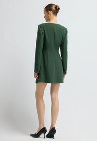 Frau mit einem Dutt, die ein maßgeschneidertes, dunkelgrünes langärmliges Minikleid und schwarze strukturierte Absatzschuhe trägt, steht vor einem einfarbigen Hintergrund.