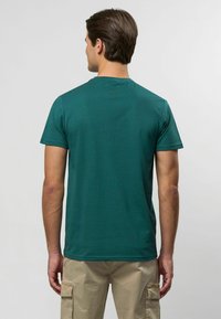 Zaļš īsām piedurknēm T-krekls, izgatavots no gludas auduma, ar apaļu izgriezumu un nelielu tekstūru. Aizmugurē redzama birku detaļa netālu no apkakles.