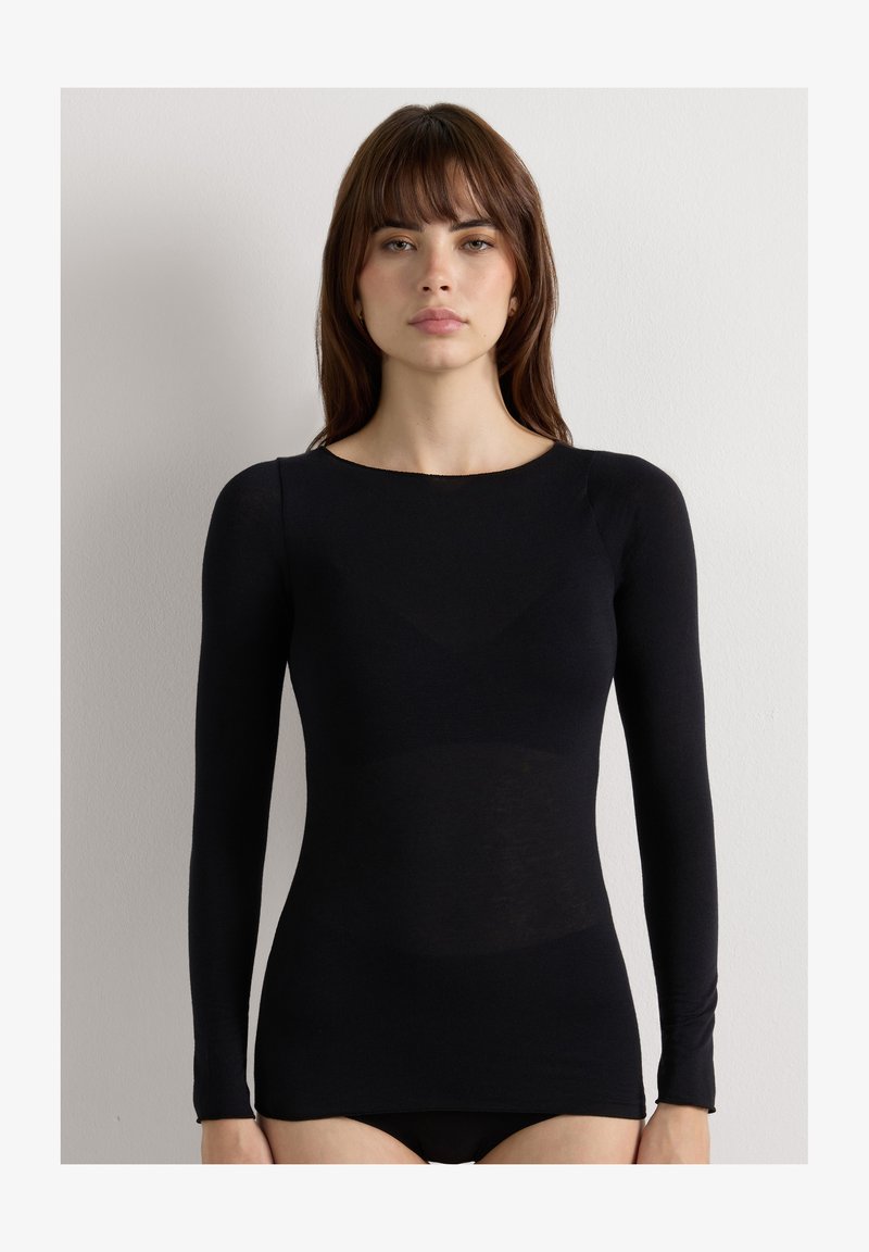 Femme aux cheveux bruns raides portant un haut noir moulant à manches longues, debout devant un fond blanc uni.