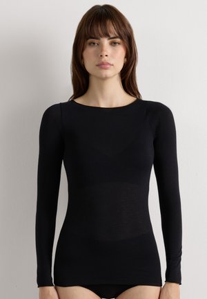 Mujer con cabello castaño liso que lleva una camiseta negra ajustada de manga larga, de pie frente a un fondo blanco liso.