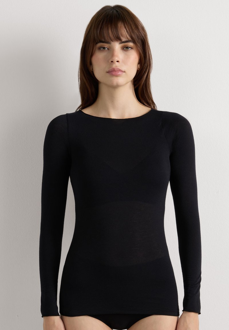 Woman with straight brown hair wearing a fitted black long-sleeve top, standing against a plain white background.