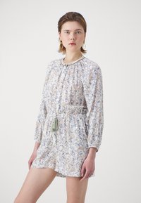 Ungdommelig kvinne iført en langt ermet blomster paisley romper med elastisk midje og dusker til snøring, stående mot en enkel hvit bakgrunn.