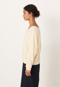 Sieviete ar cirtainiem matiem pītā bulciņā, valkā krēmkrāsas džemperi bez pleciem un tumšu svītrainu svārku, stāv sānis skats uz vienkārša fona.