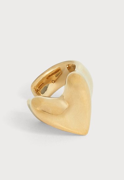 Une bague sculpturale de couleur dorée, au large motif incurvé en forme de cœur, présentée sur un fond blanc uni.