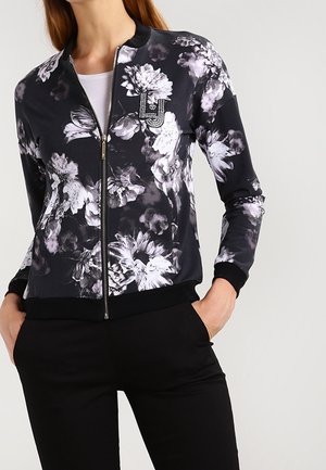 Woman wearing a black zip-up jacket with white and gray floral patterns, a small "LJ" logo on the chest, and black pants with hands in pockets.