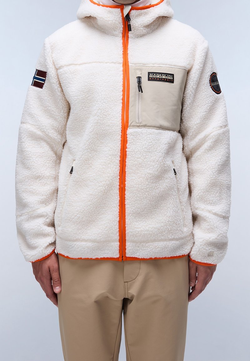Man som bär en vit fleecejacka med orangea detaljer, beige bröstficka och norsk flagga patch på vänster arm.