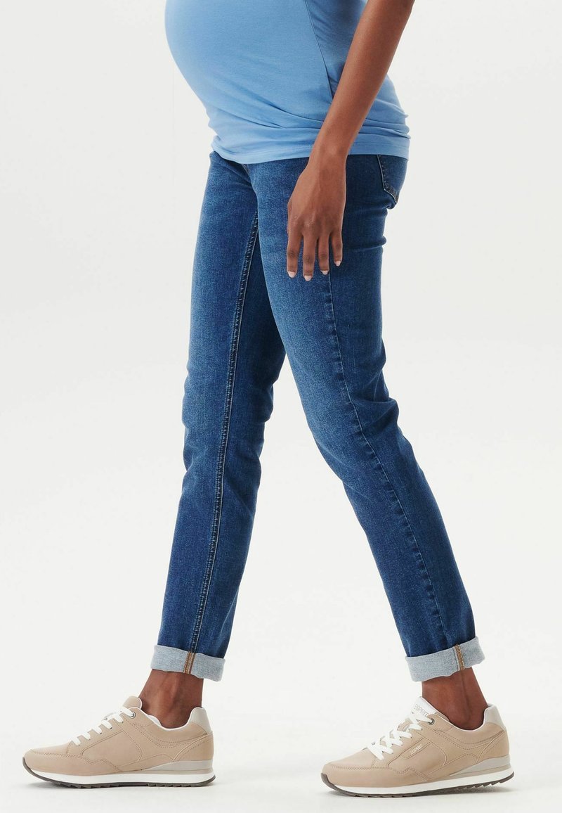 Graviditets skinny jeans i mörkblå denim med en slät yta, med uppvikta fållar. Kombinerat med ljusbeige sneakers.