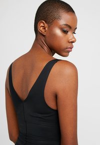 Μαύρο αμάνικο τοπ με σχέδιο V στην πλάτη. Το ύφασμα είναι λείος και εφαρμοστός, τονίζοντας τις καμπύλες του σώματος.